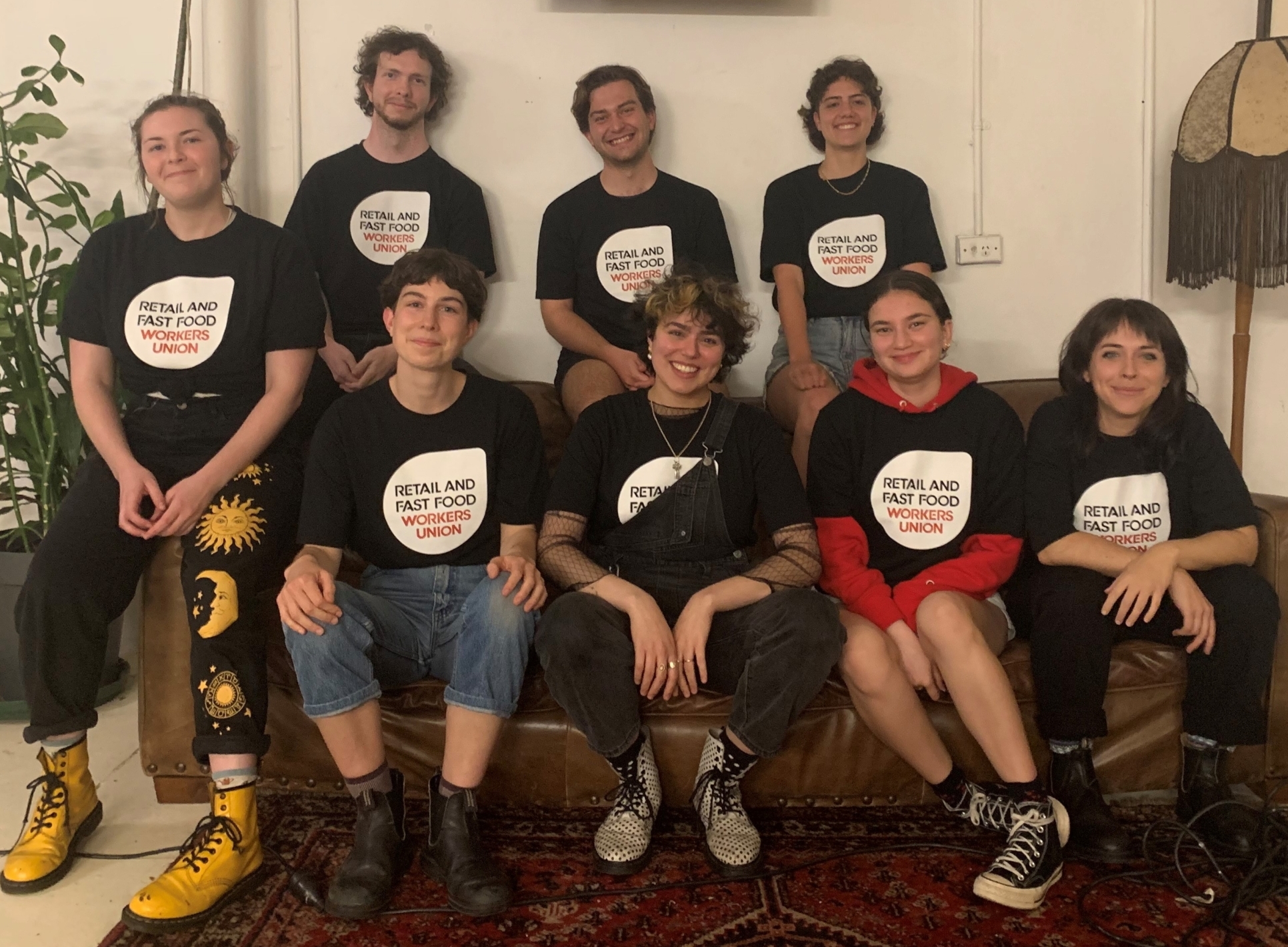A group photograph of eight workers and union members from the bookstore Better Read Than Dead. Four are seated on a couch and four are standing behind. All are wearing black t-shirts with the Retail and Fast Food Workers Union logo in white, red and black.