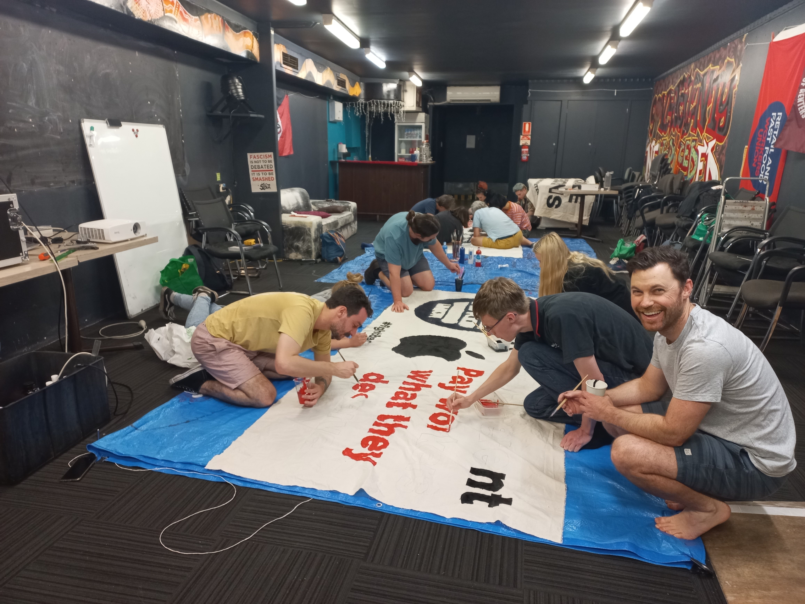A photograph of a union banner being painted by Apple workers, the worker closest to the camera is smiling.