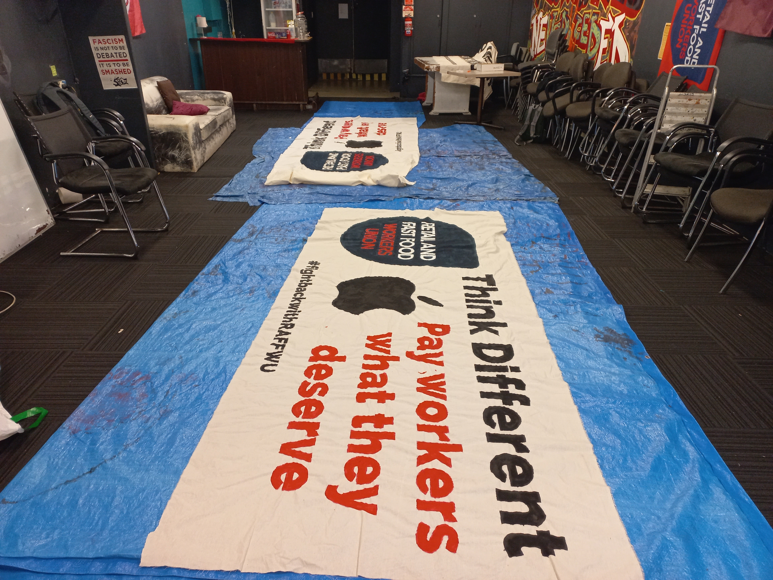 A photograph of a banner that has the Apple logo on it and the slogan "Think Different, Pay workers what they deserve".